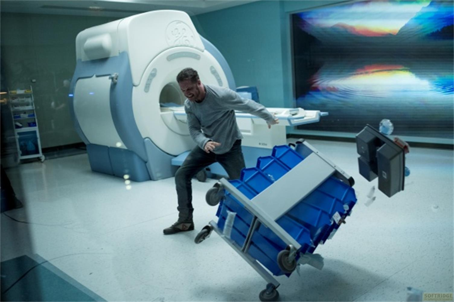 Uomo inciampa su carrello blu davanti a macchina MRI. Oggetti volano.