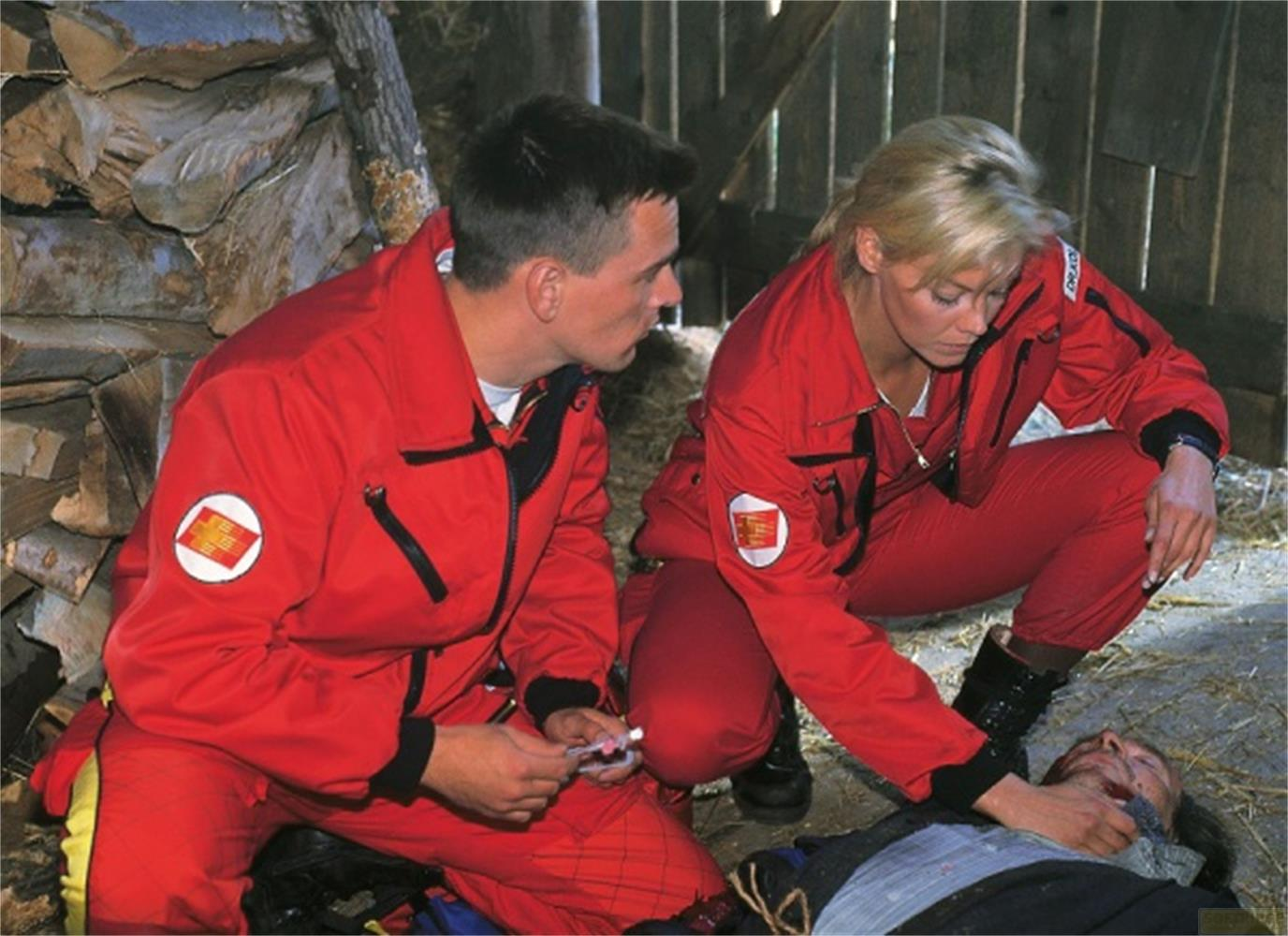 Due paramedici in rosso esaminano un uomo sdraiato. Legna da ardere sullo sfondo.