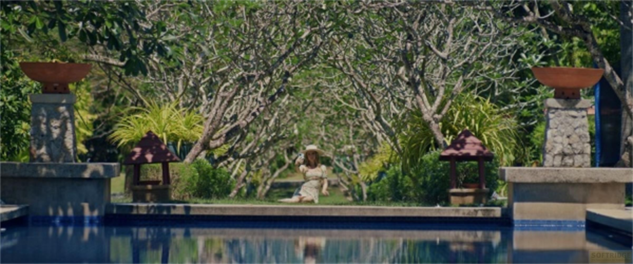 Una donna con cappello siede sul bordo della piscina, con alberi e cespugli dietro.