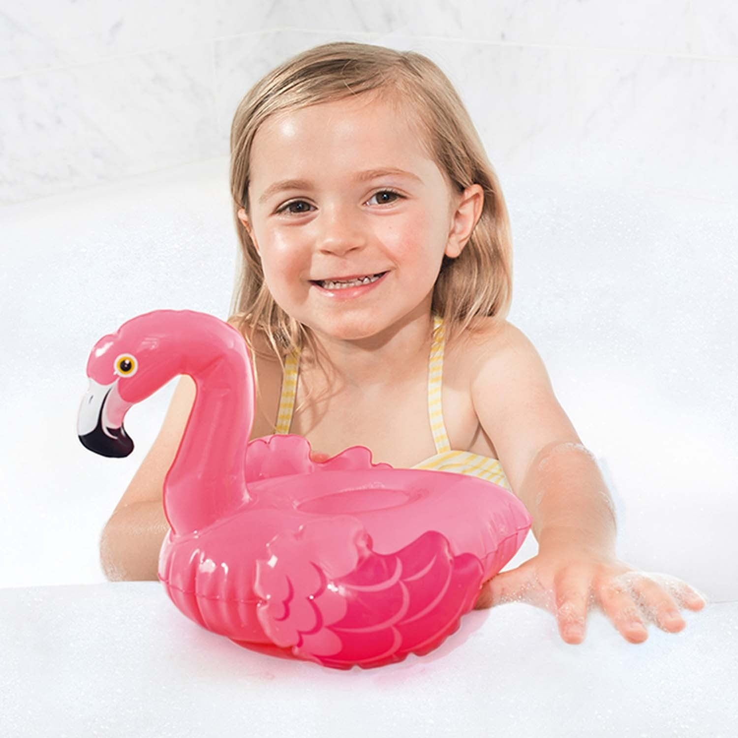 Una niña sonriente con traje de baño amarillo sostiene un flotador de flamenco rosa en una bañera burbujeante.