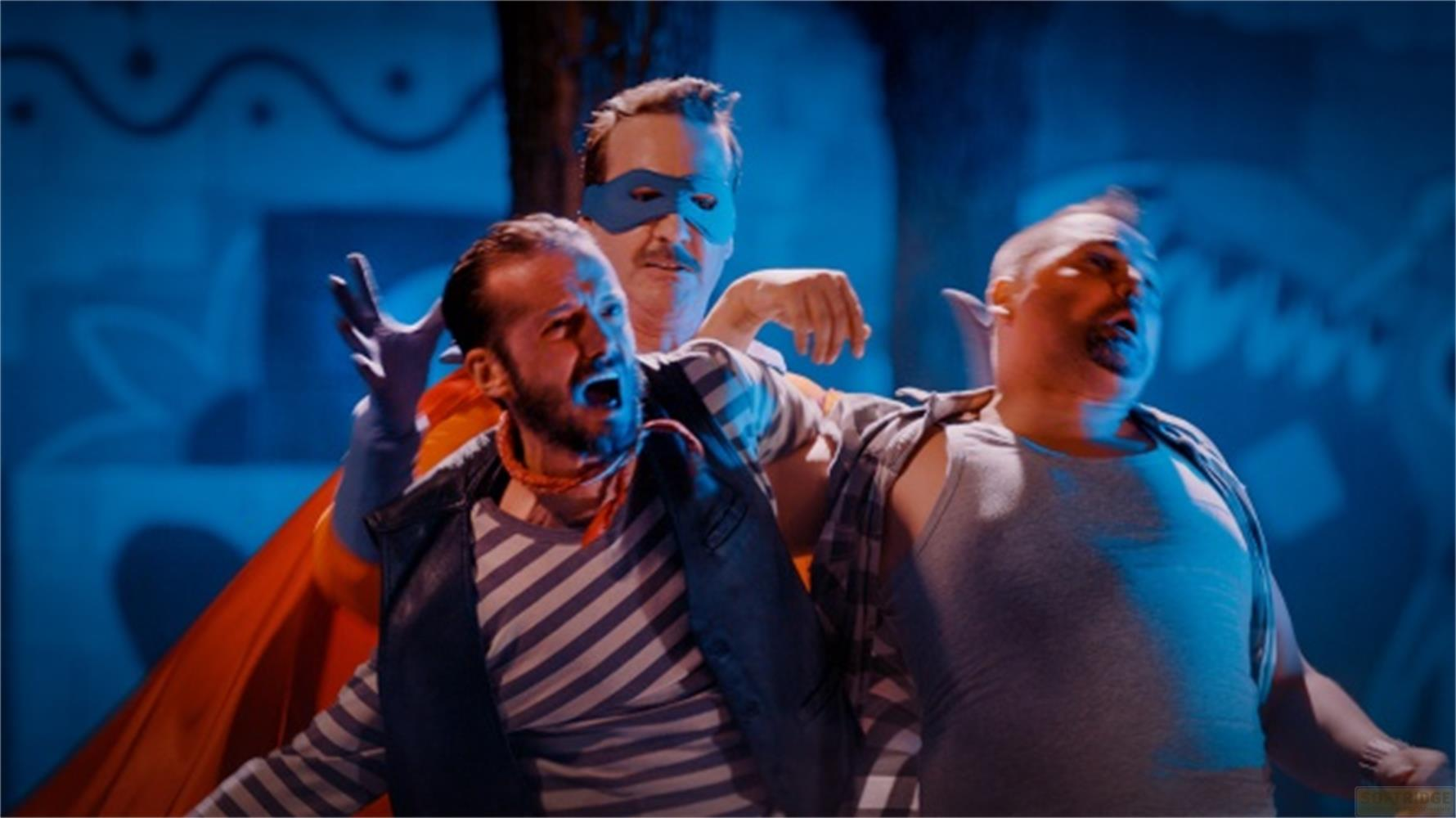 Tre uomini in costume, uno mascherato, urlano su sfondo blu.