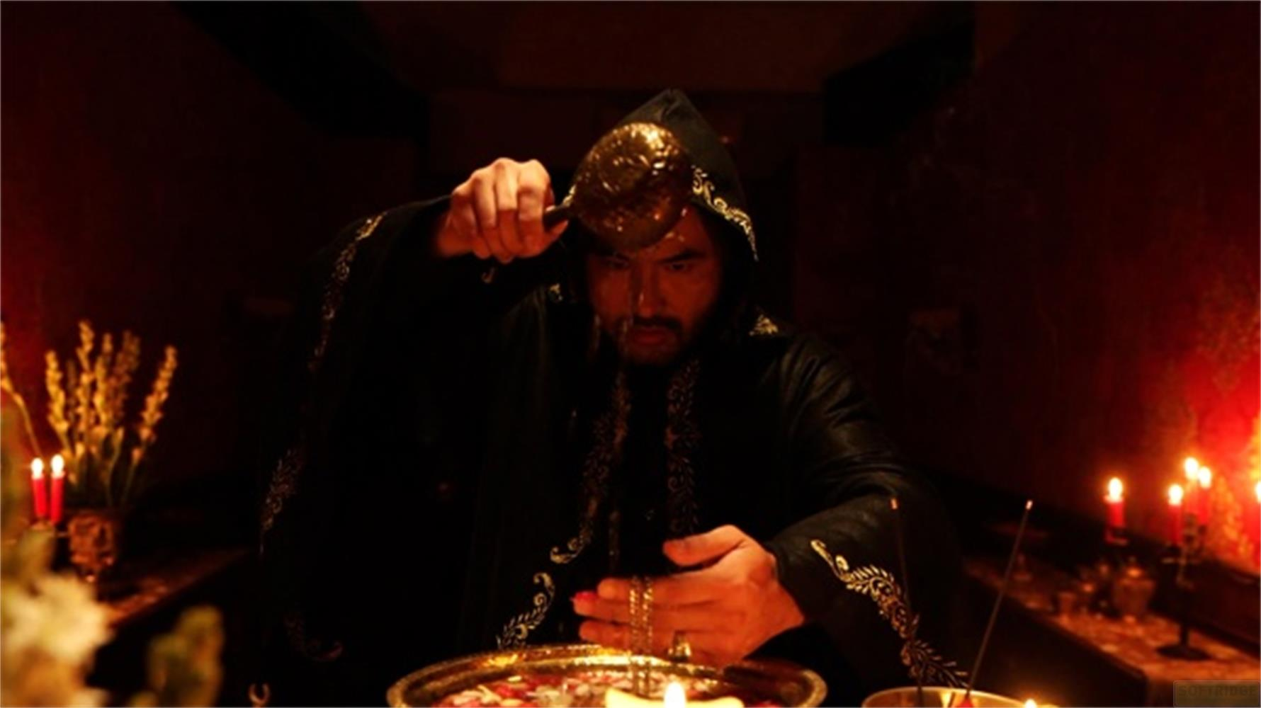 Uomo in veste con cappuccio versa liquido da coppa dorata in bacino, circondato da candele.