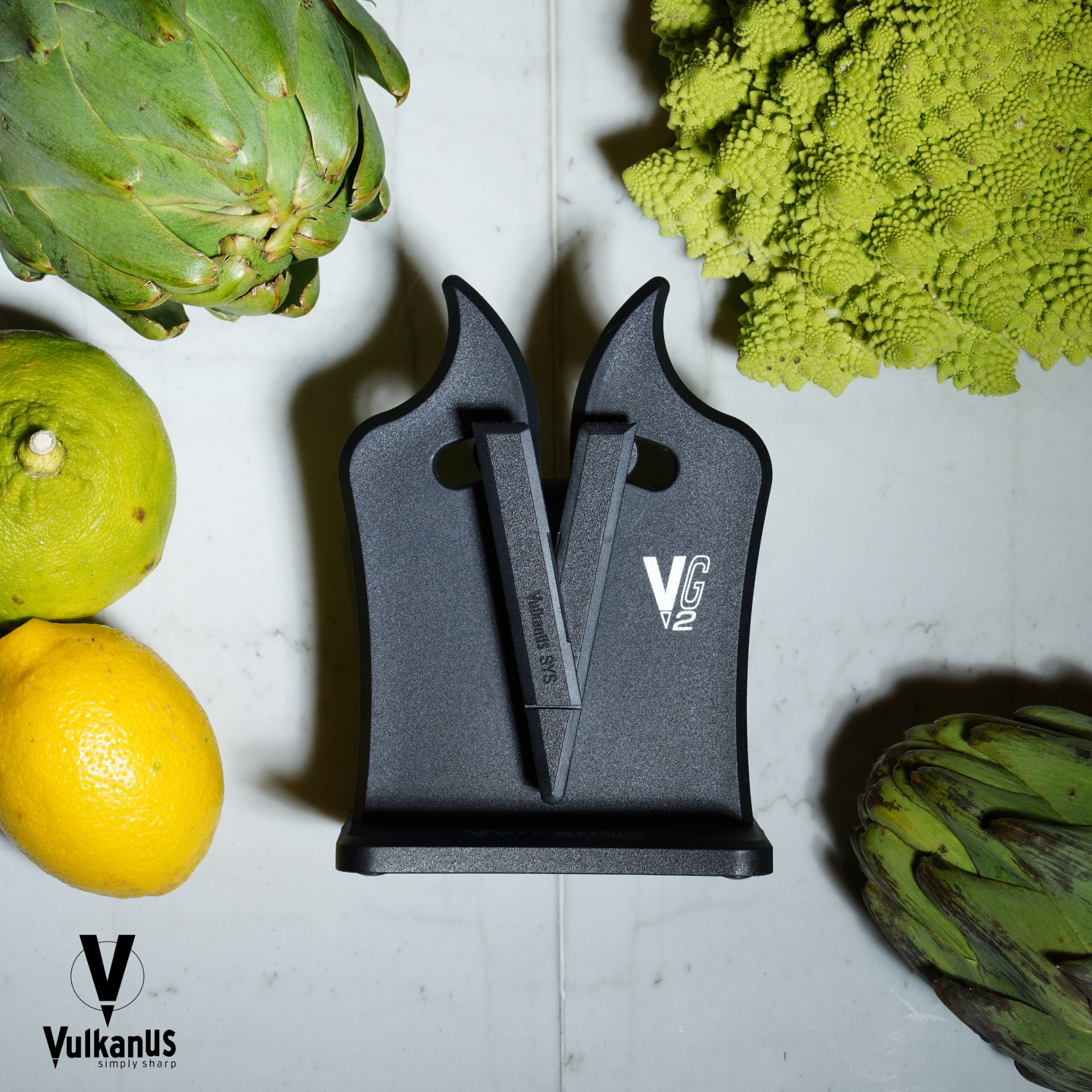 Un aiguiseur de couteaux noir est posé entre des artichauts, un citron, une lime et du Romanesco.