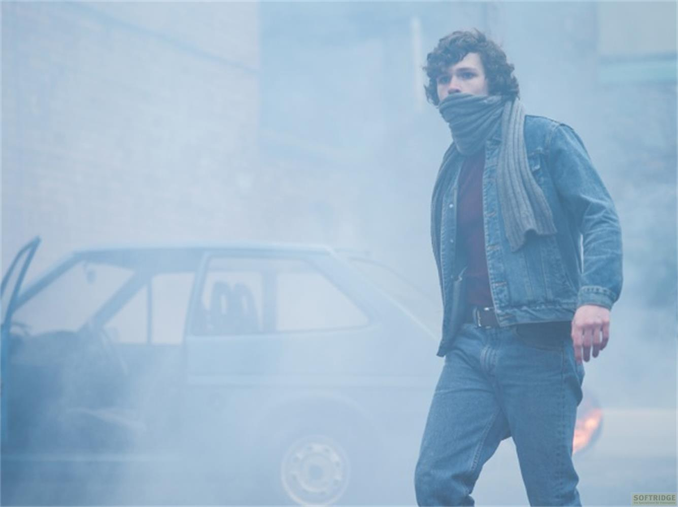 Uomo in giacca di jeans e sciarpa passa accanto a un'auto, avvolto dalla nebbia.