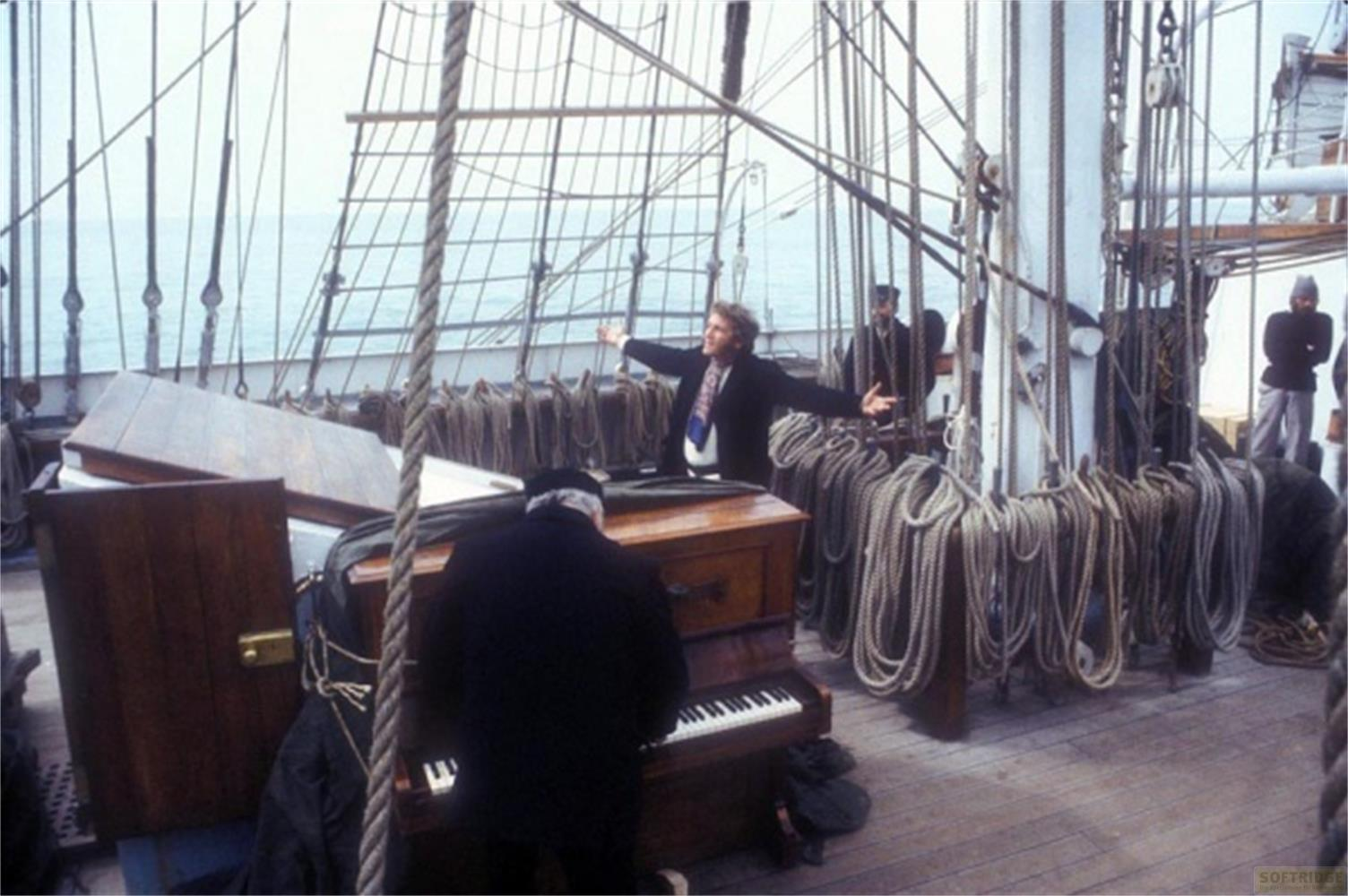 Un uomo suona il piano su una nave mentre un altro canta a braccia aperte.