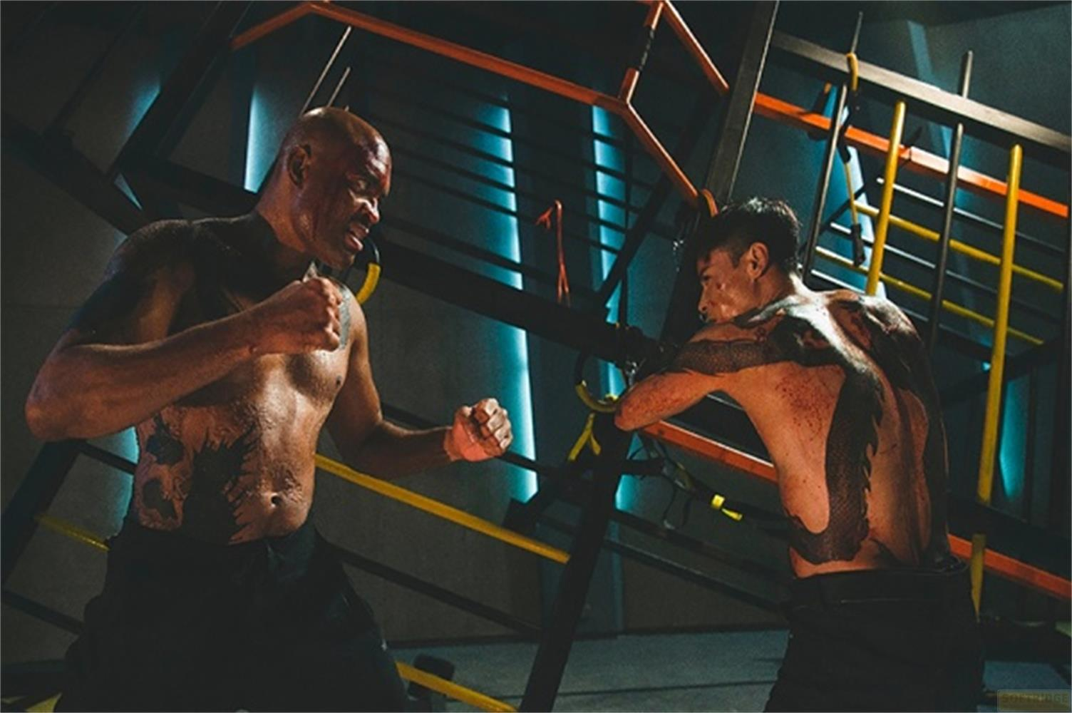 Due uomini tatuati combattono in una palestra con attrezzature per esercizi.