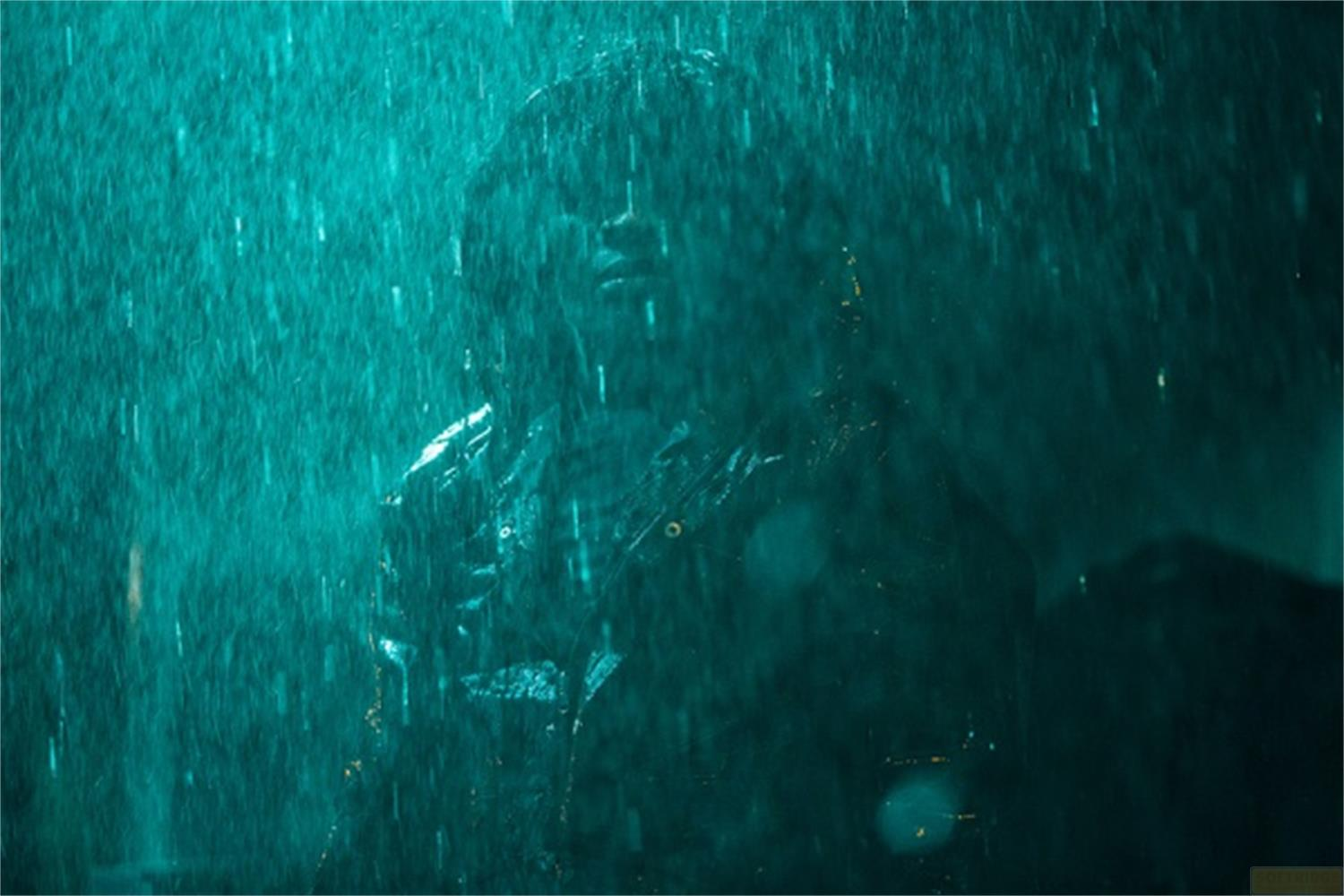 Una persona è in piedi sotto una pioggia battente, illuminata da una luce verde acqua minacciosa.