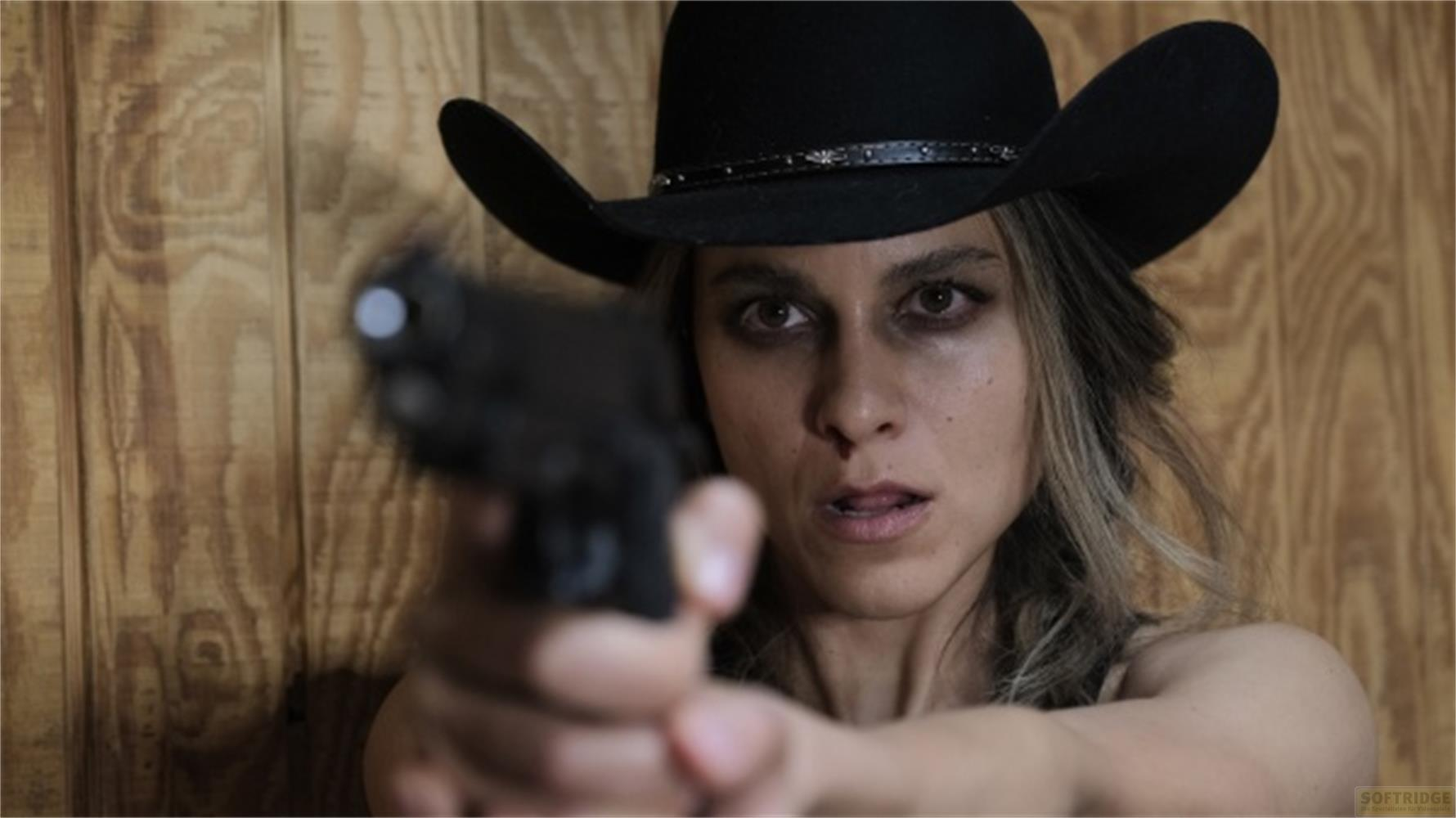Una donna con cappello da cowboy punta una pistola davanti a un muro di legno.