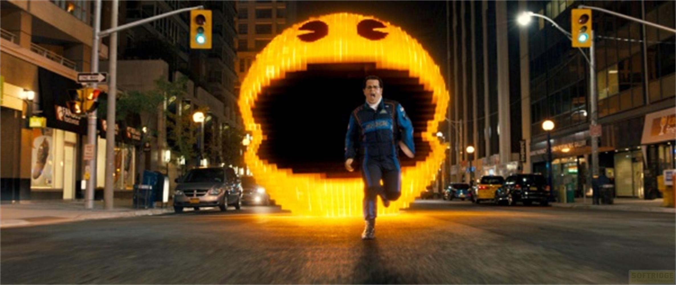 Ein Mann in einem blauen Anzug rennt nachts auf einer Stadtstraße vor einem riesigen, leuchtenden Pac-Man.