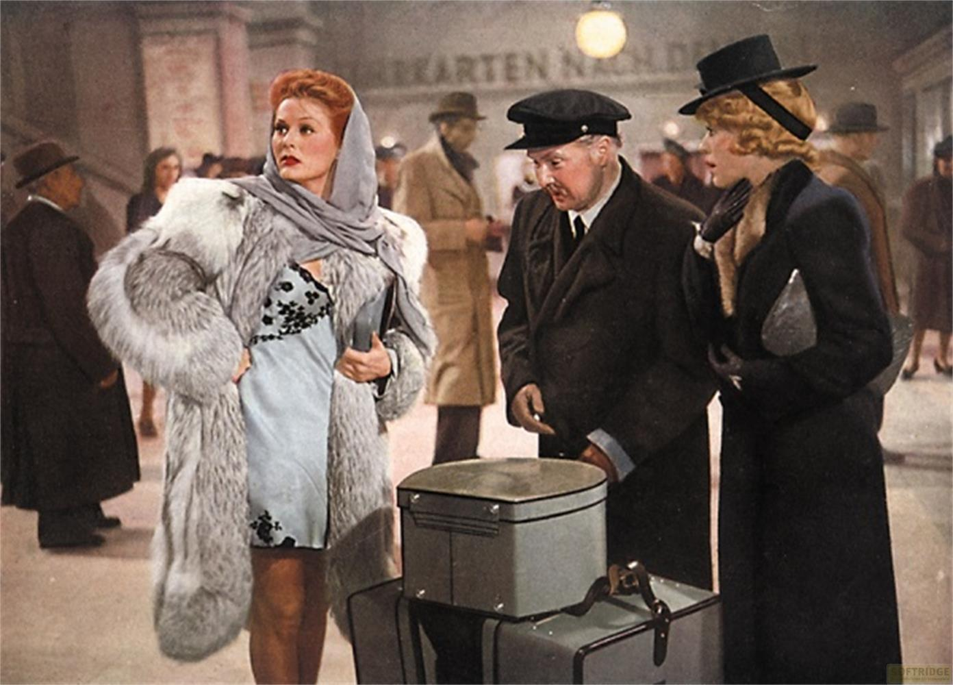 Donna in pelliccia, sottoveste e foulard sta con bagagli. Persone dietro.