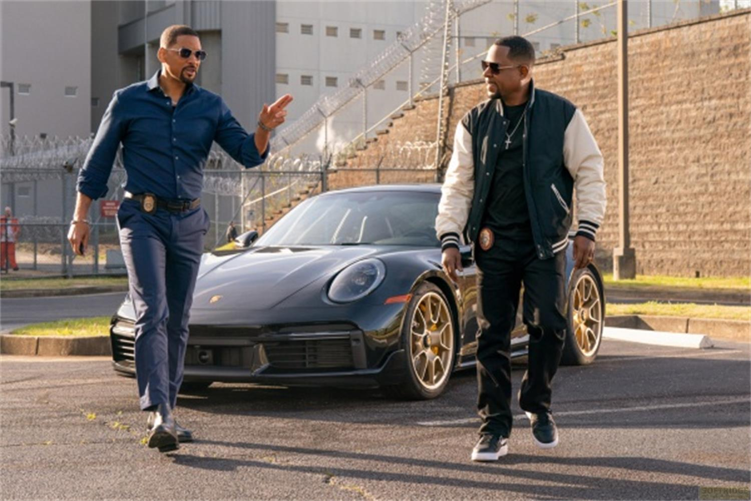 Due uomini camminano accanto a una Porsche nera. Uno indossa un abito blu, l'altro una giacca bomber.
