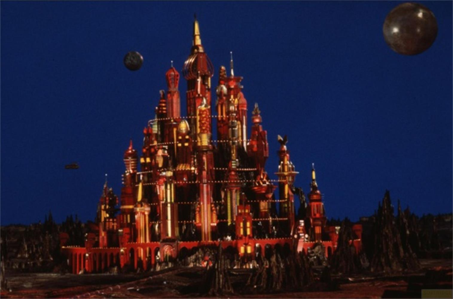 Un castello rosso e illuminato con guglie su un paesaggio scuro e roccioso sotto un cielo blu.