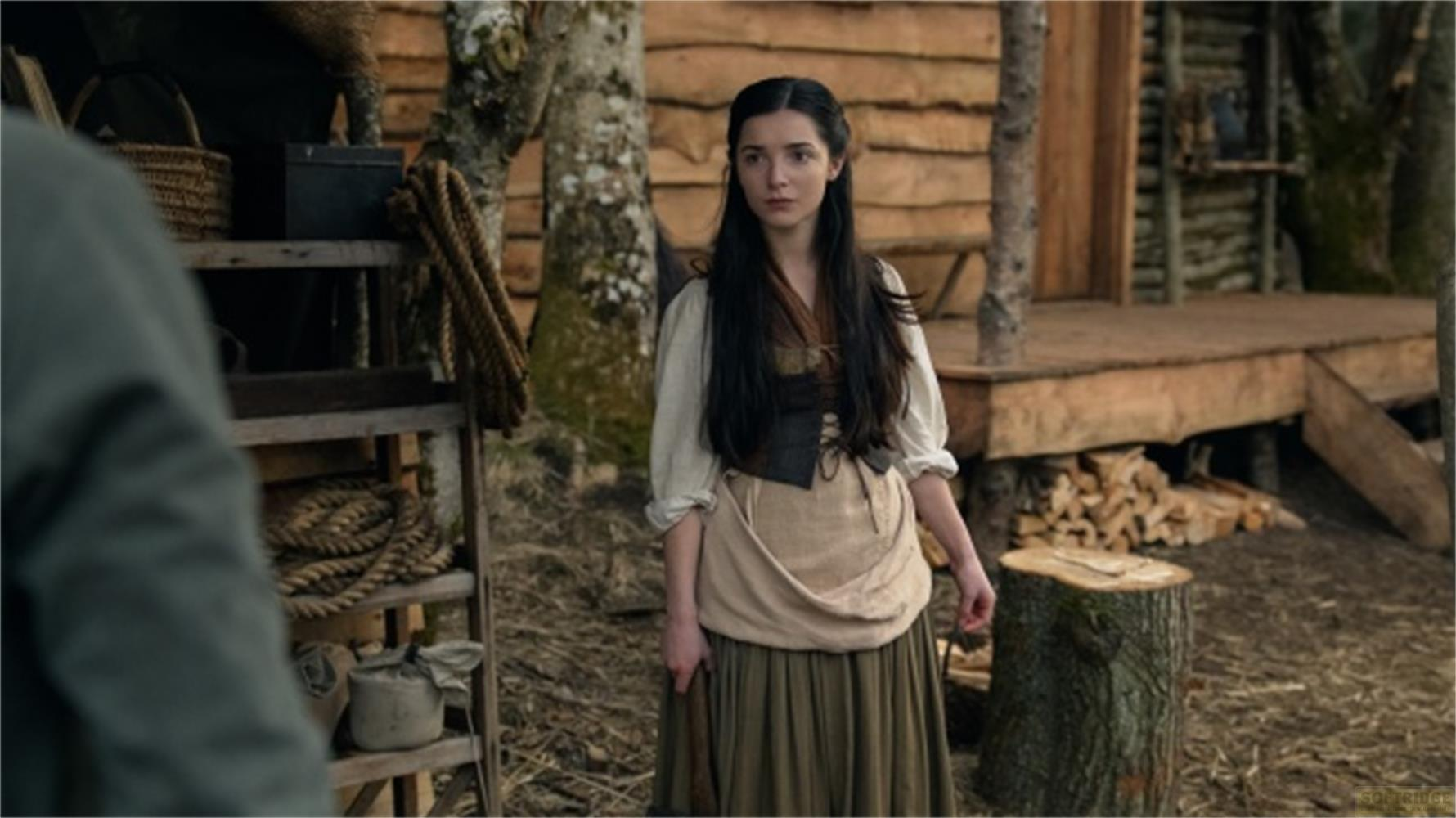 Junge Frau in historischer Kleidung steht vor einem Holzhaus mit Korb und Seilen.