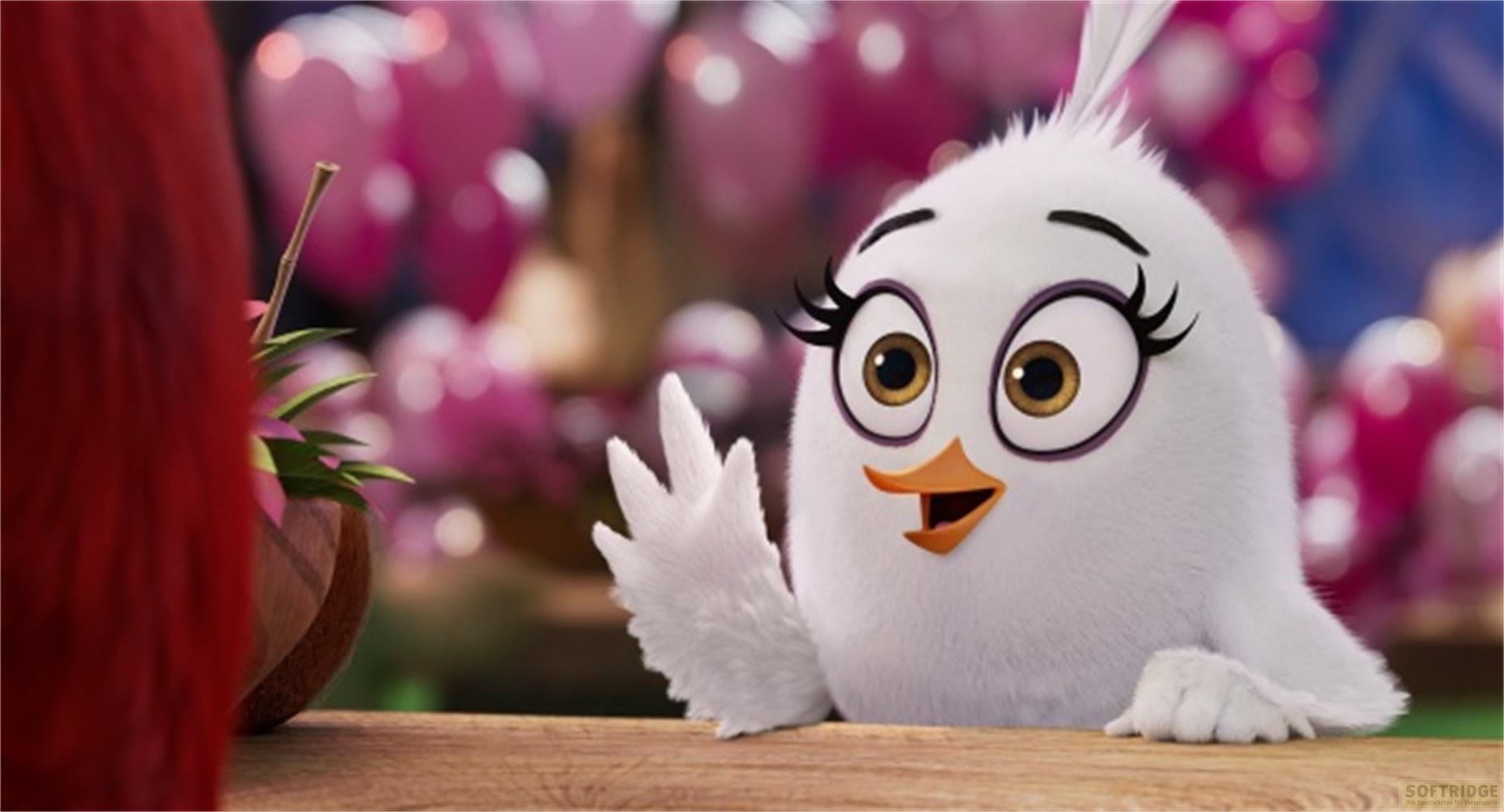 Eine weiße Vogelfigur winkt mit der Pfote, mit pinken Ballons im Hintergrund.