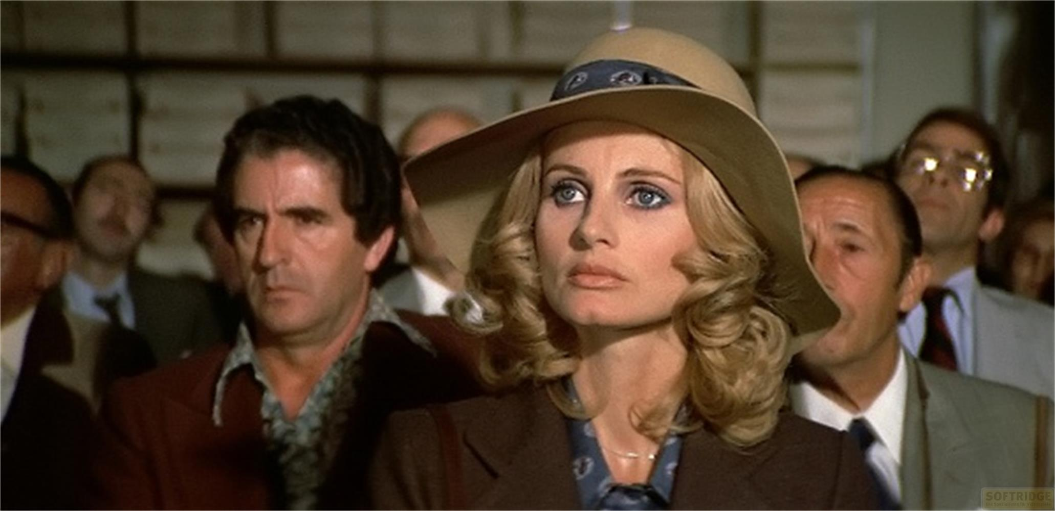 Une femme au chapeau et boucles blondes regarde en l'air, des hommes au fond.