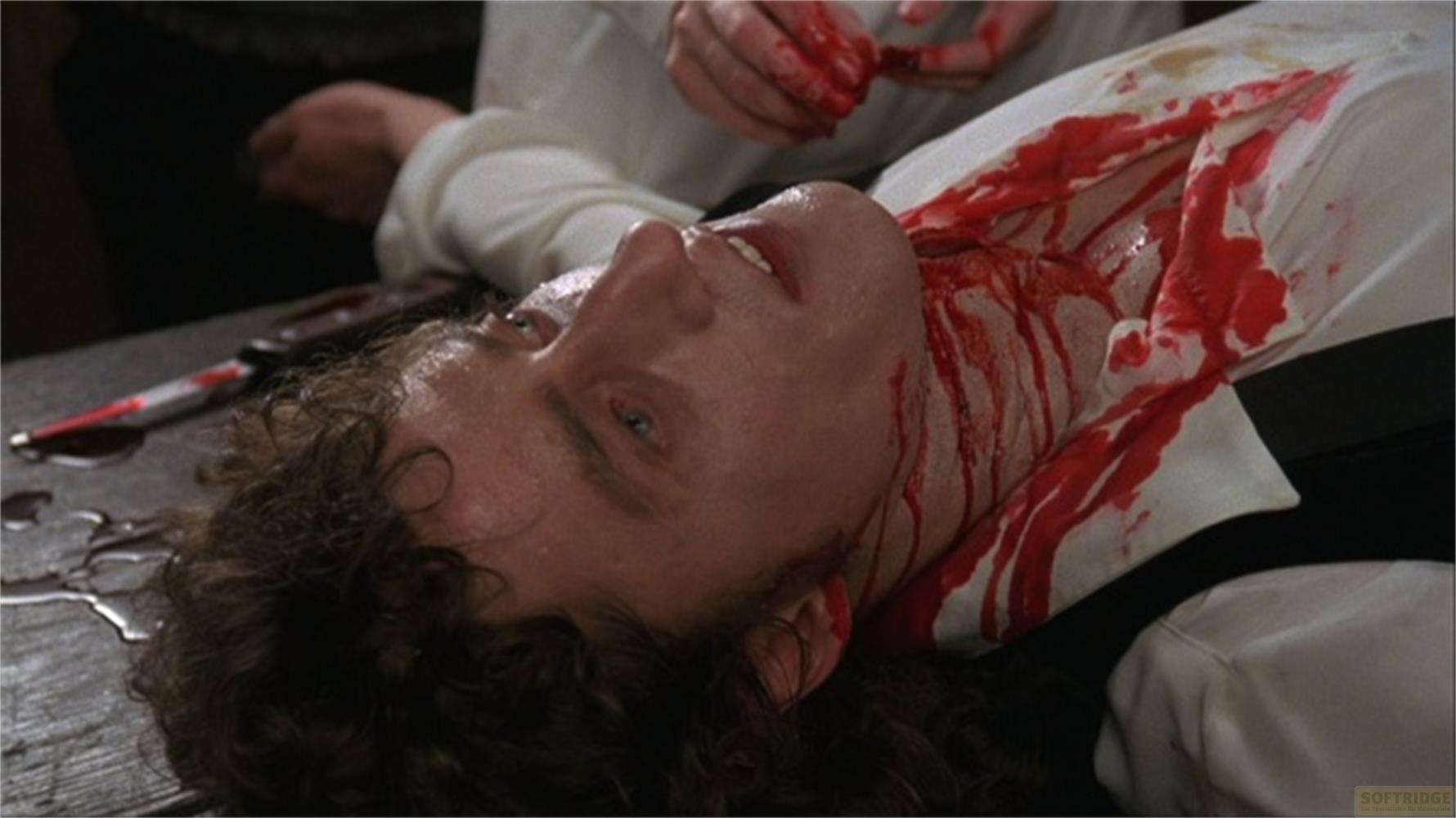 Un uomo giace su un tavolo, coperto di sangue, con un coltello accanto.