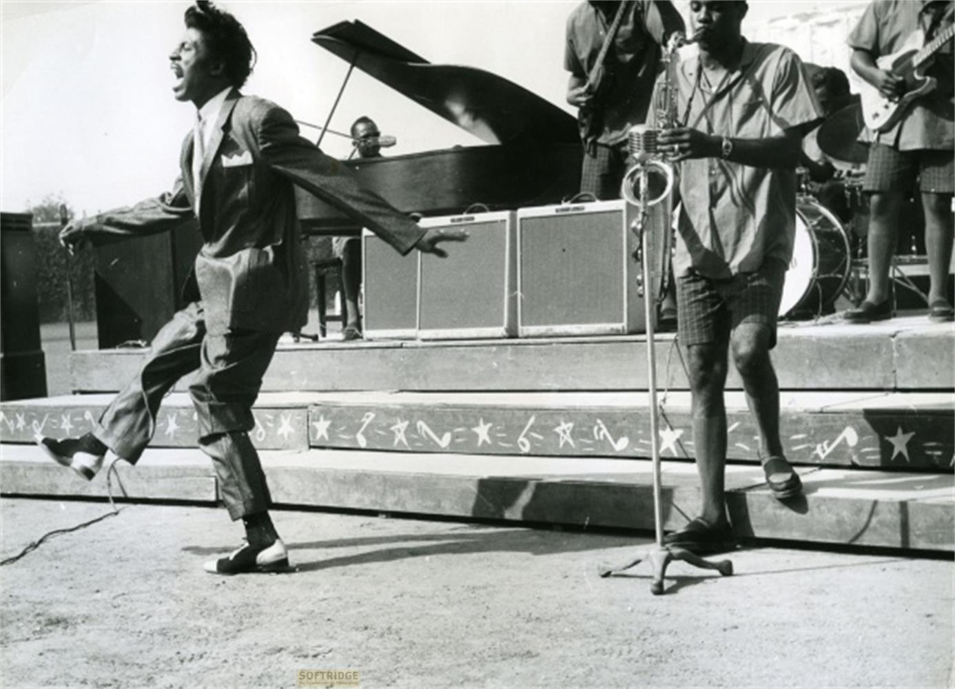 Un uomo in abito danza sul palco mentre una band suona. È visibile un pianoforte.