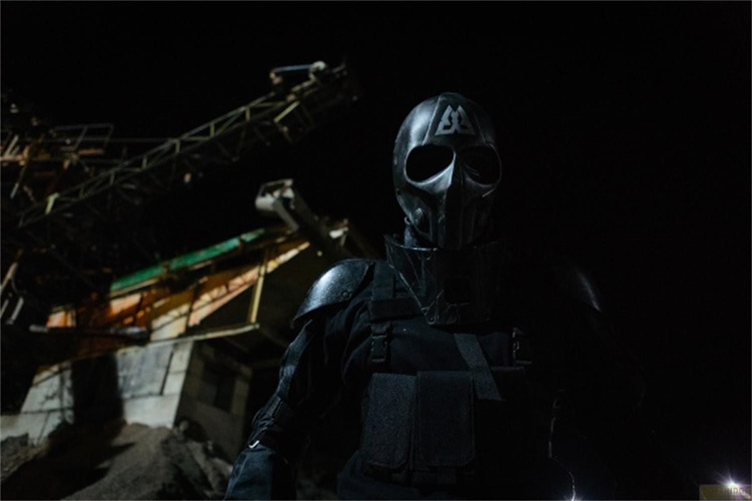 Una persona in armatura e maschera scure sta davanti a un edificio industriale di notte.