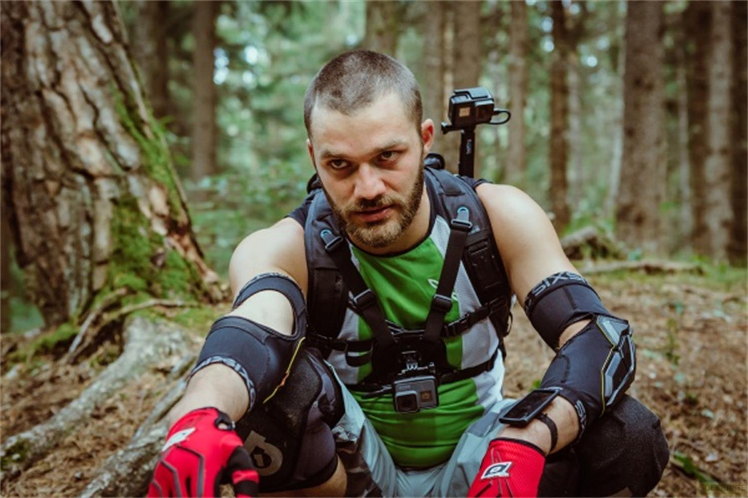 Un uomo con barba, equipaggiamento protettivo e fotocamera siede in un bosco.