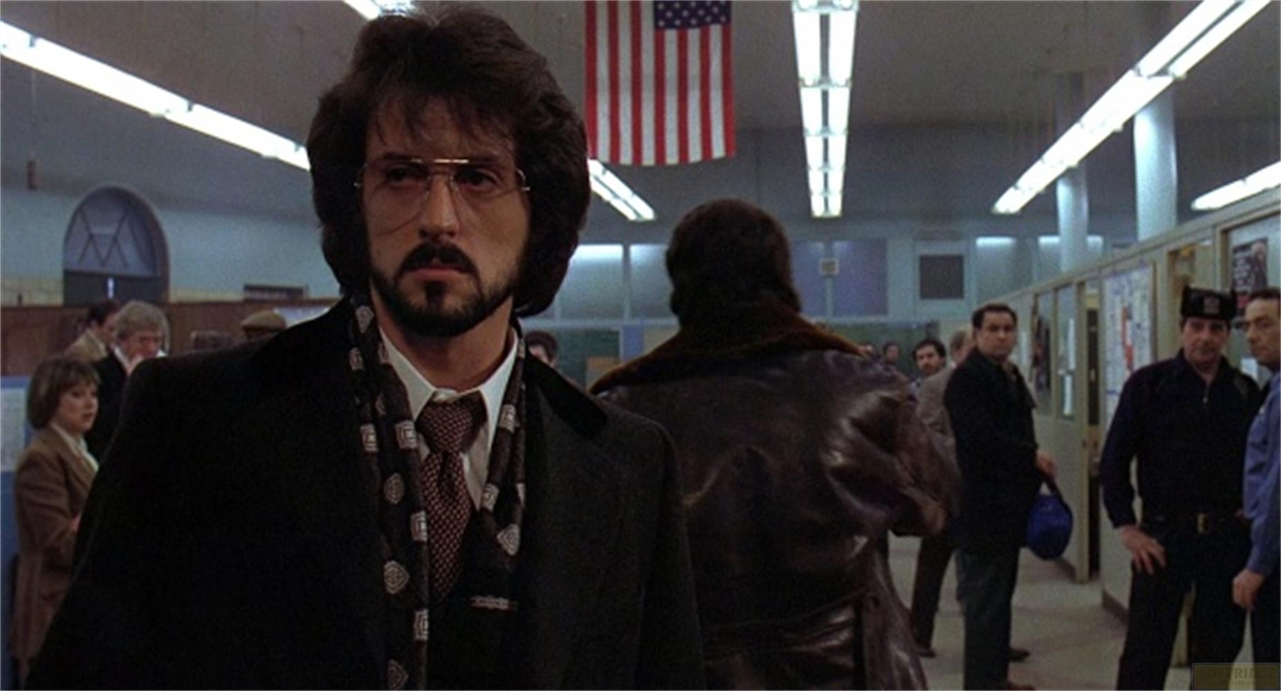 Uomo con occhiali e barba davanti a bandiera USA. Altre persone sullo sfondo.
