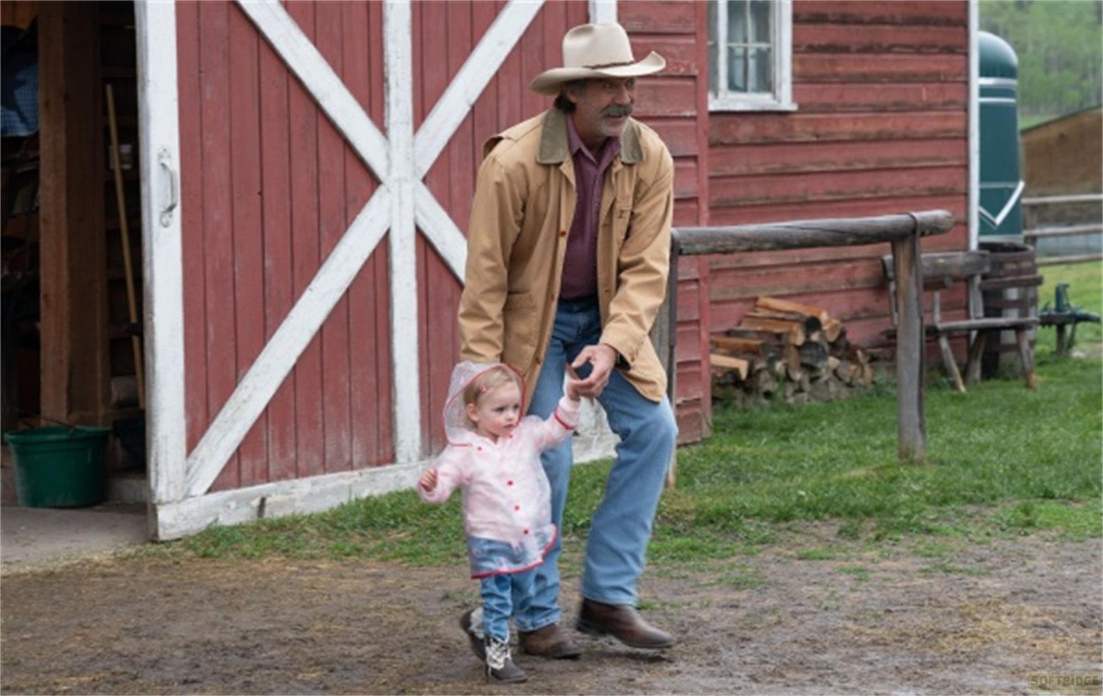 Un uomo con cappello da cowboy tiene per mano un bambino davanti a un fienile.
