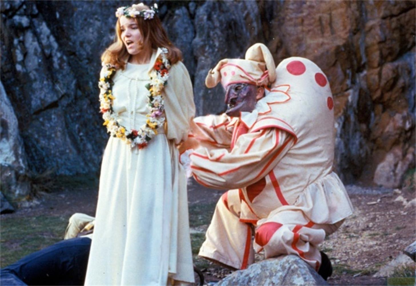 Una donna in abito bianco e corona di fiori parla a una figura in costume da clown.