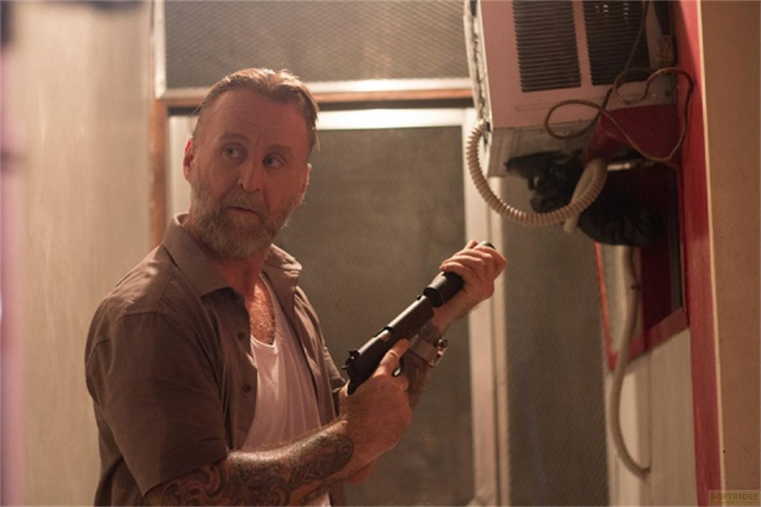 Un uomo con barba e tatuaggi tiene una pistola vicino a un condizionatore.