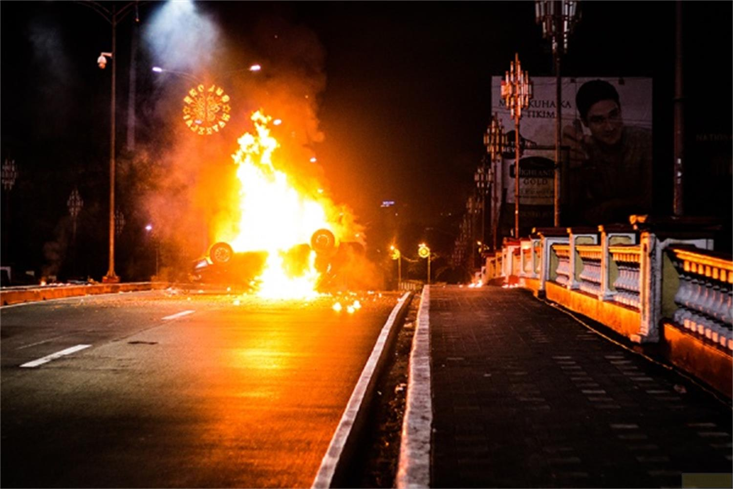 Un'auto in fiamme su una strada buia di notte, con fumo e fiamme.