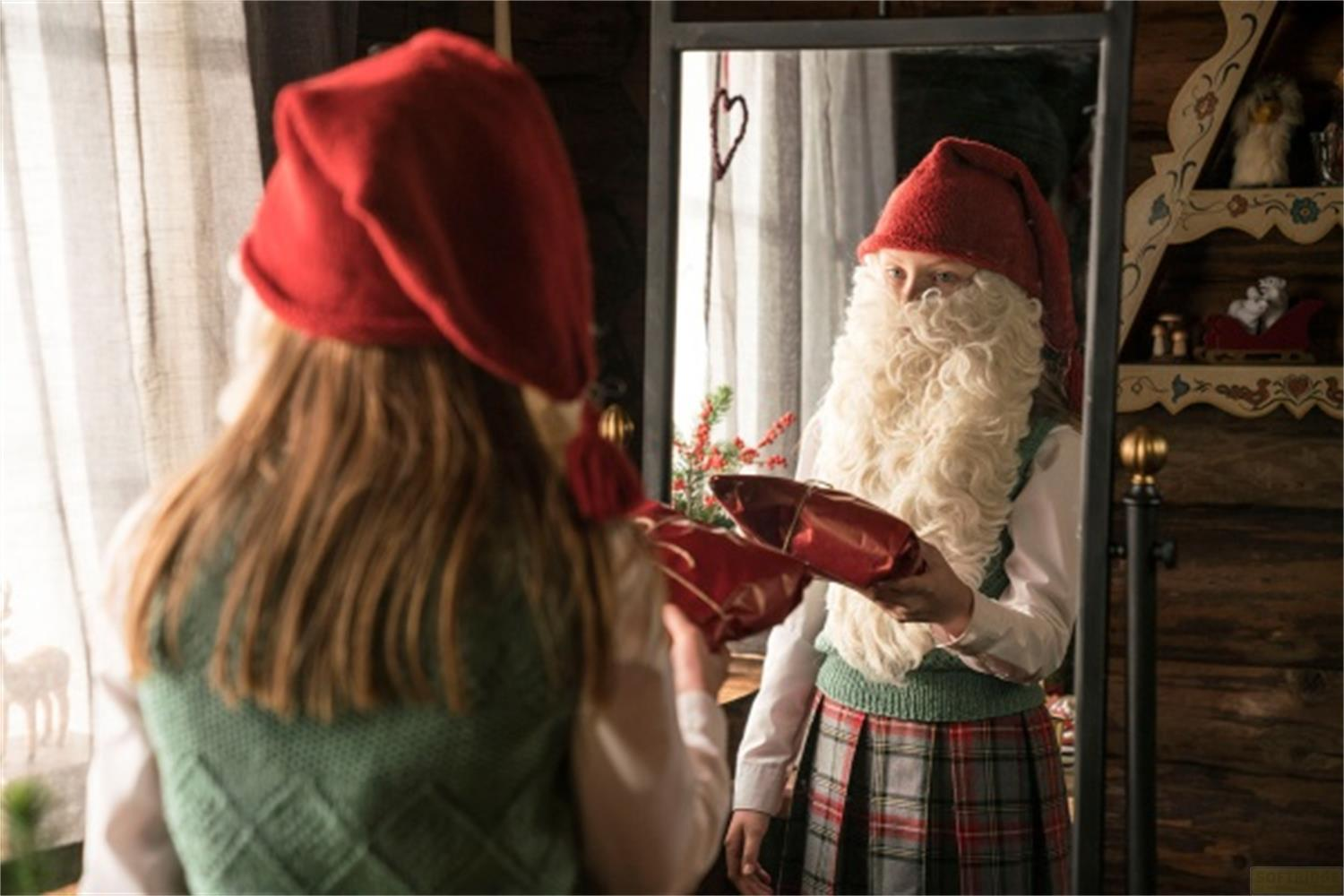 Enfant avec barbe de Père Noël et bonnet rouge tient des cadeaux, reflété dans un miroir.