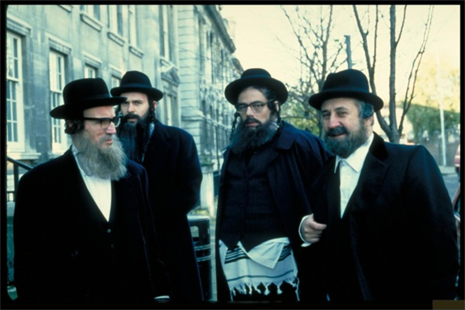 Quattro uomini con cappelli neri e abiti camminano all'aperto. Tre hanno la barba, uno porta gli occhiali.