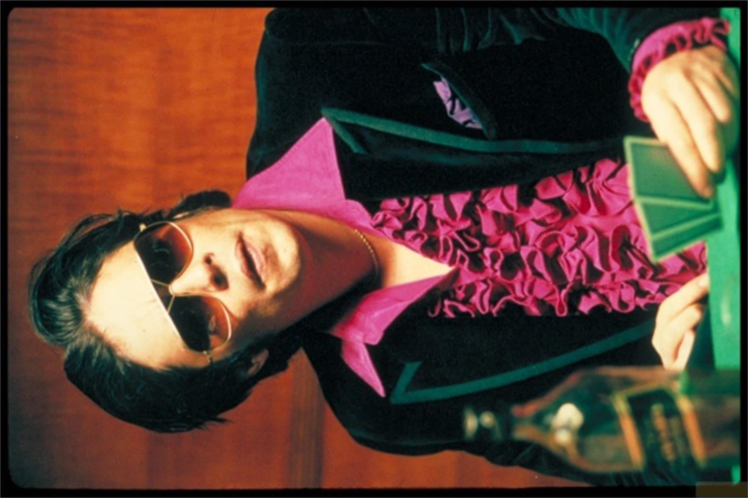 Persona con occhiali da sole e camicia con volant tiene in mano delle carte.
