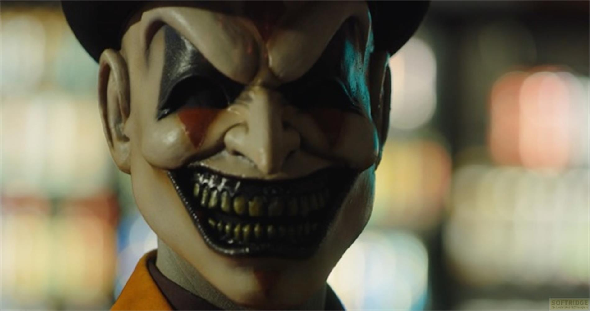Primo piano di una maschera da clown inquietante con cappello nero, denti gialli e giacca arancione.