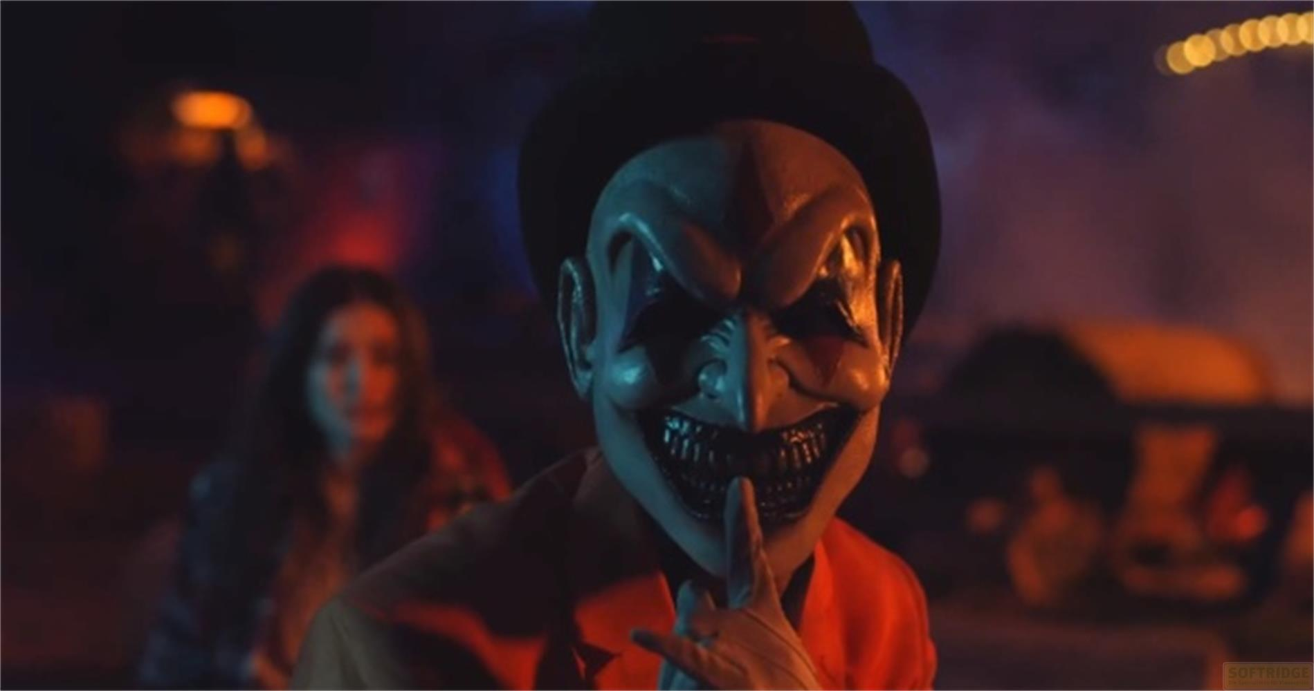 Un uomo con maschera da clown e cappello nero porta un dito alla bocca. Una donna sullo sfondo.