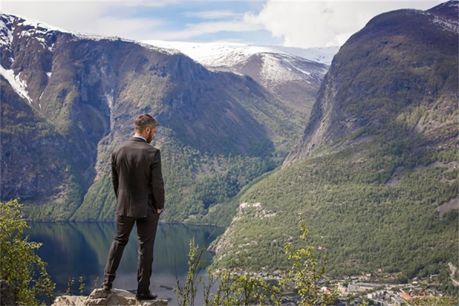 Homme en costume sur des rochers surplombant un lac et des montagnes.