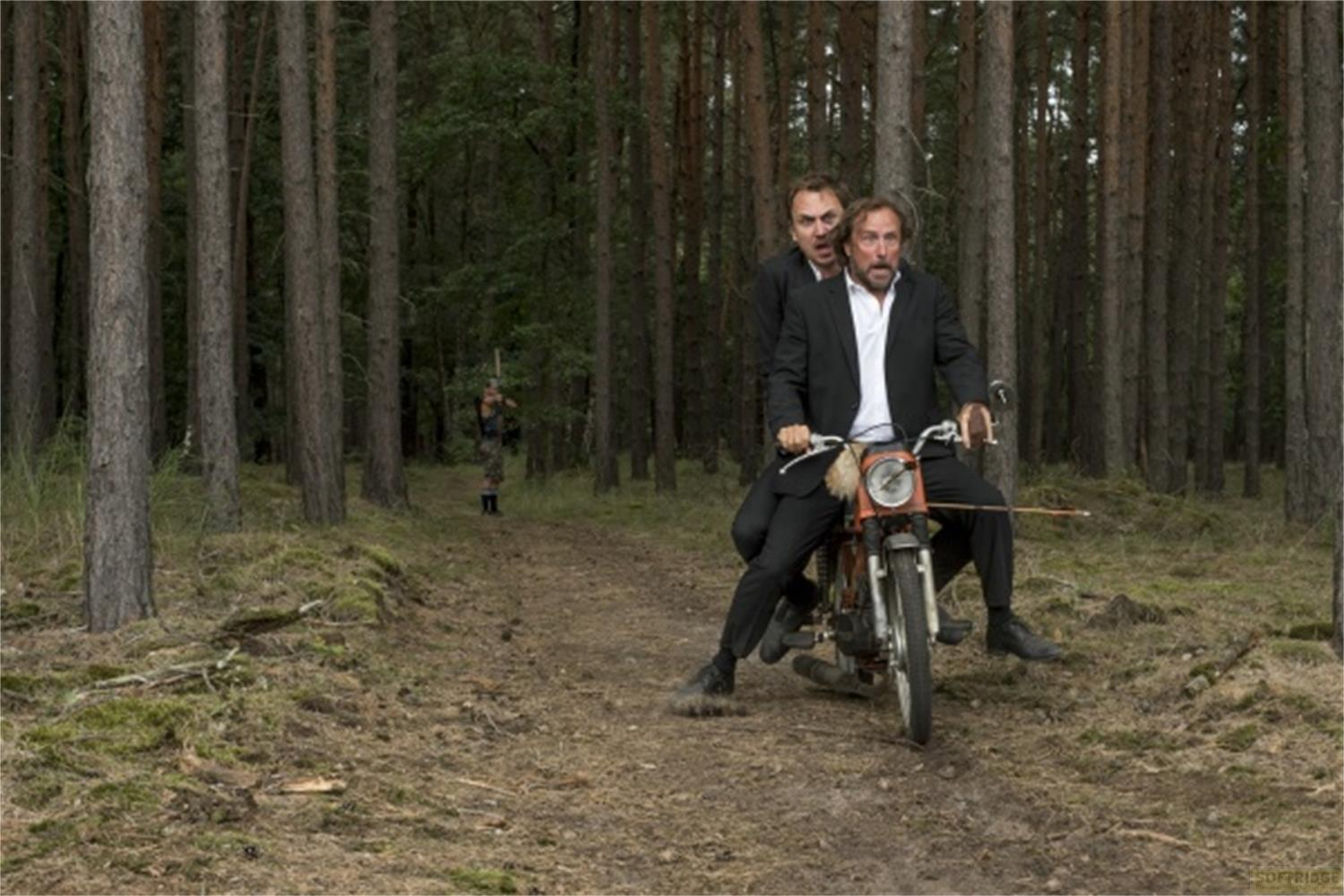 Zwei Männer auf einem Motorrad im Wald, ein Mann im Hintergrund.