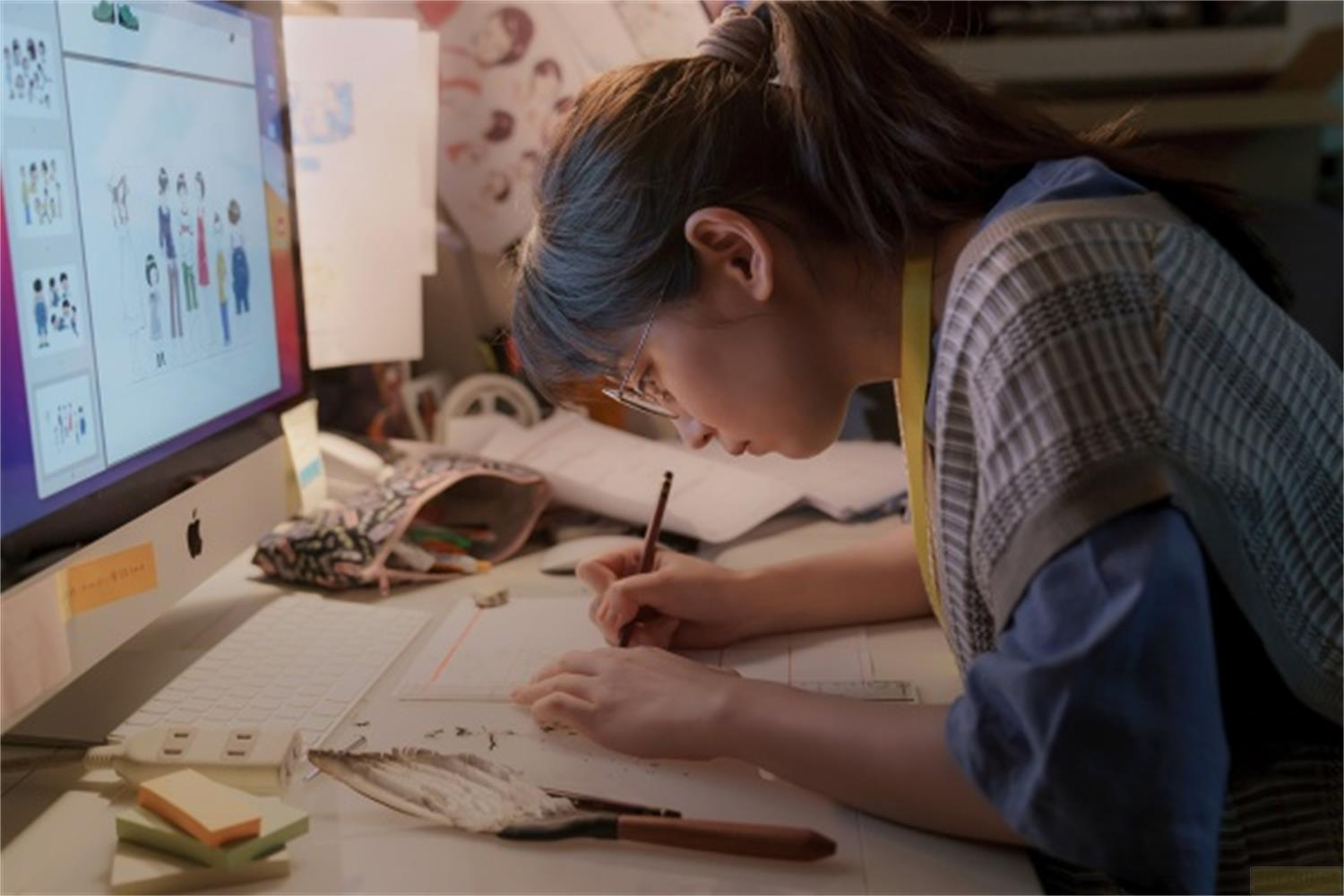 Una donna disegna a una scrivania, con un computer sullo sfondo.