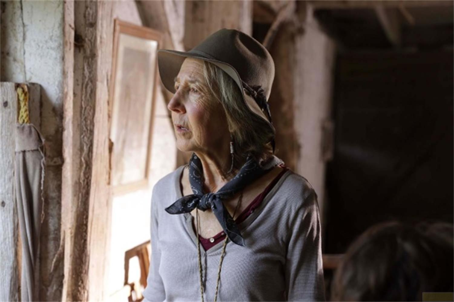 Donna anziana con cappello e foulard guarda fuori dalla finestra in ambiente rustico.