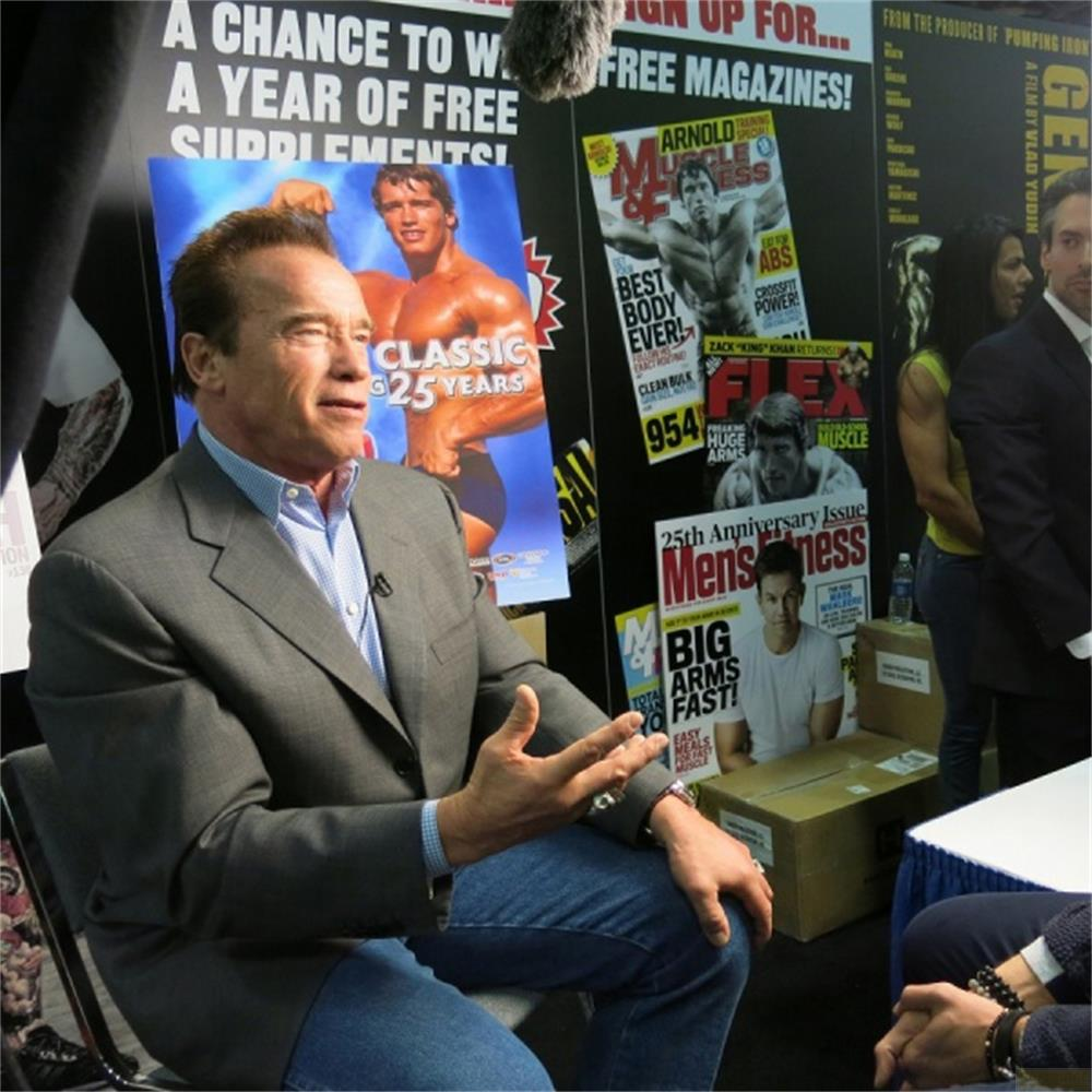 Arnold Schwarzenegger sitzt in einem Anzug vor Postern von Fitnessmagazinen und sich selbst.