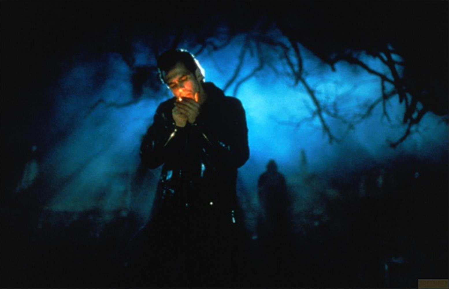 Un homme en veste sombre allume une cigarette dans une scène extérieure brumeuse et éclairée en bleu, avec des silhouettes et des arbres.