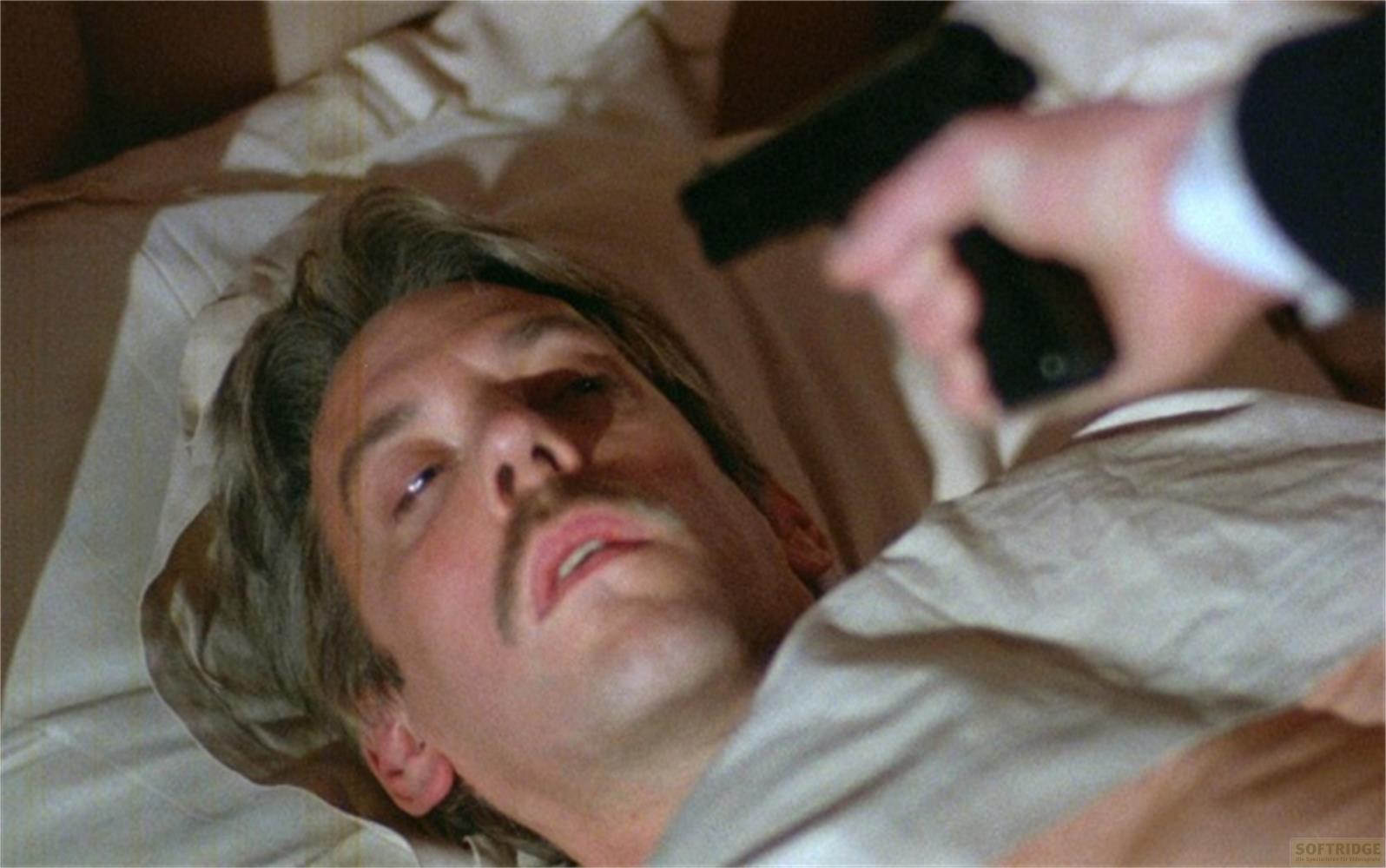 Un uomo giace a letto mentre una pistola è puntata su di lui.
