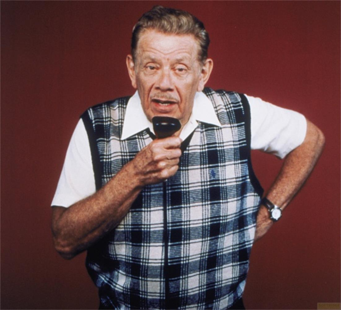Uomo anziano con microfono, indossa un gilet a quadri su camicia bianca.