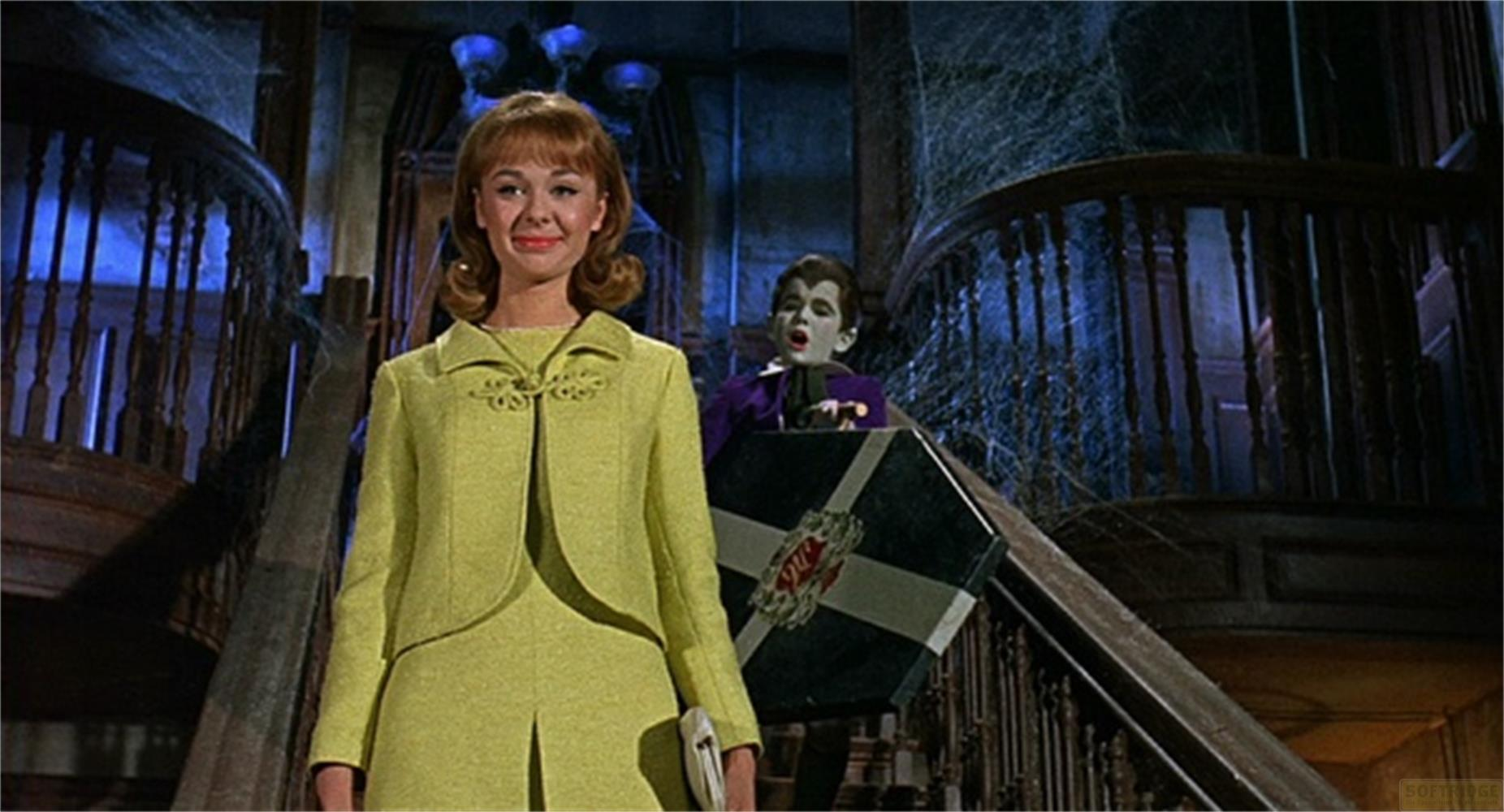 Una donna in tailleur giallo in primo piano, un bambino vampiro viola con una bara sullo sfondo.