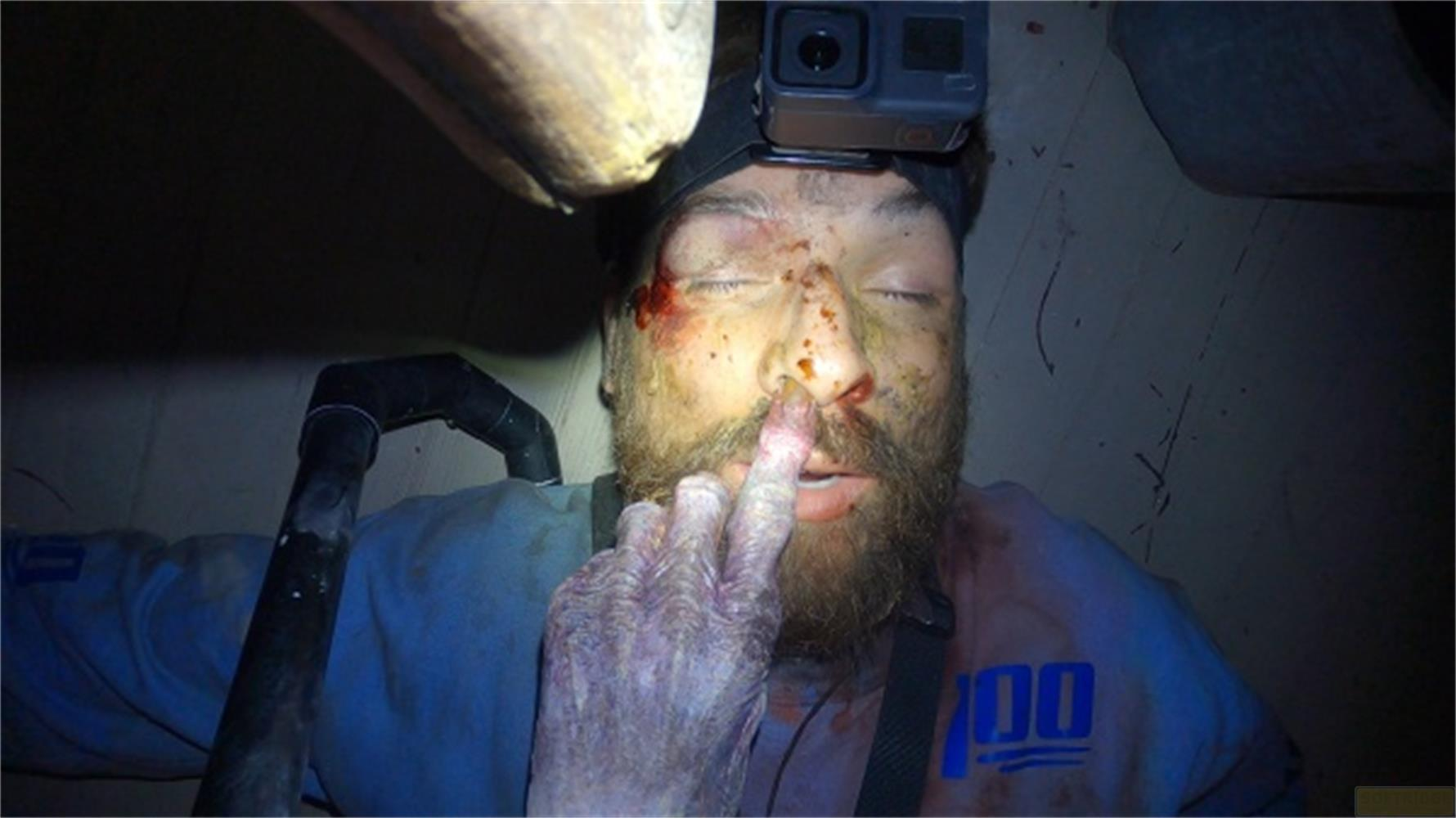 Un uomo con camicia blu, GoPro in testa, sporco e sangue sul viso.