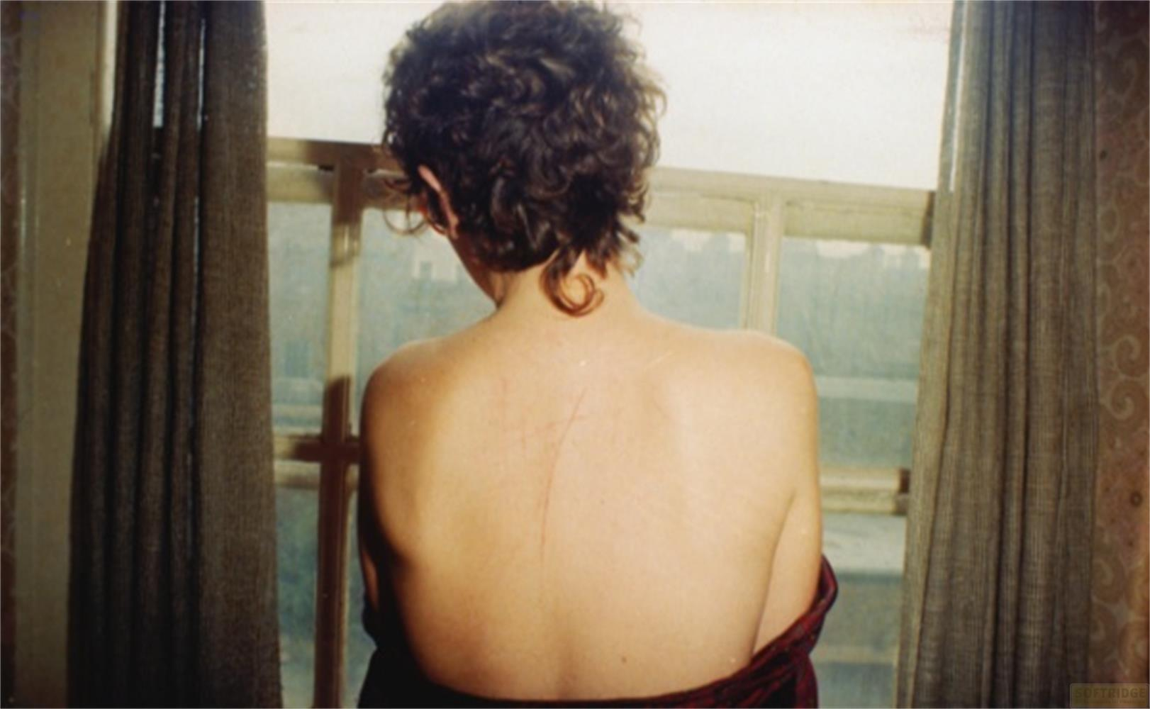 Vista posteriore di una donna con capelli corti e ricci, vicino a una finestra con tende.