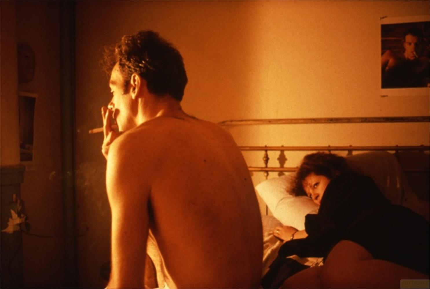Uomo fuma a letto, donna accanto. Un quadro al muro.