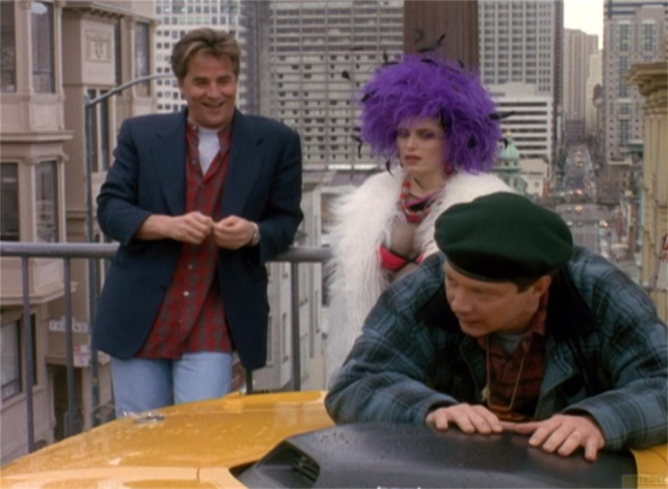 Trois personnes sur un balcon avec une ligne d'horizon urbaine. Une femme porte une perruque violette.