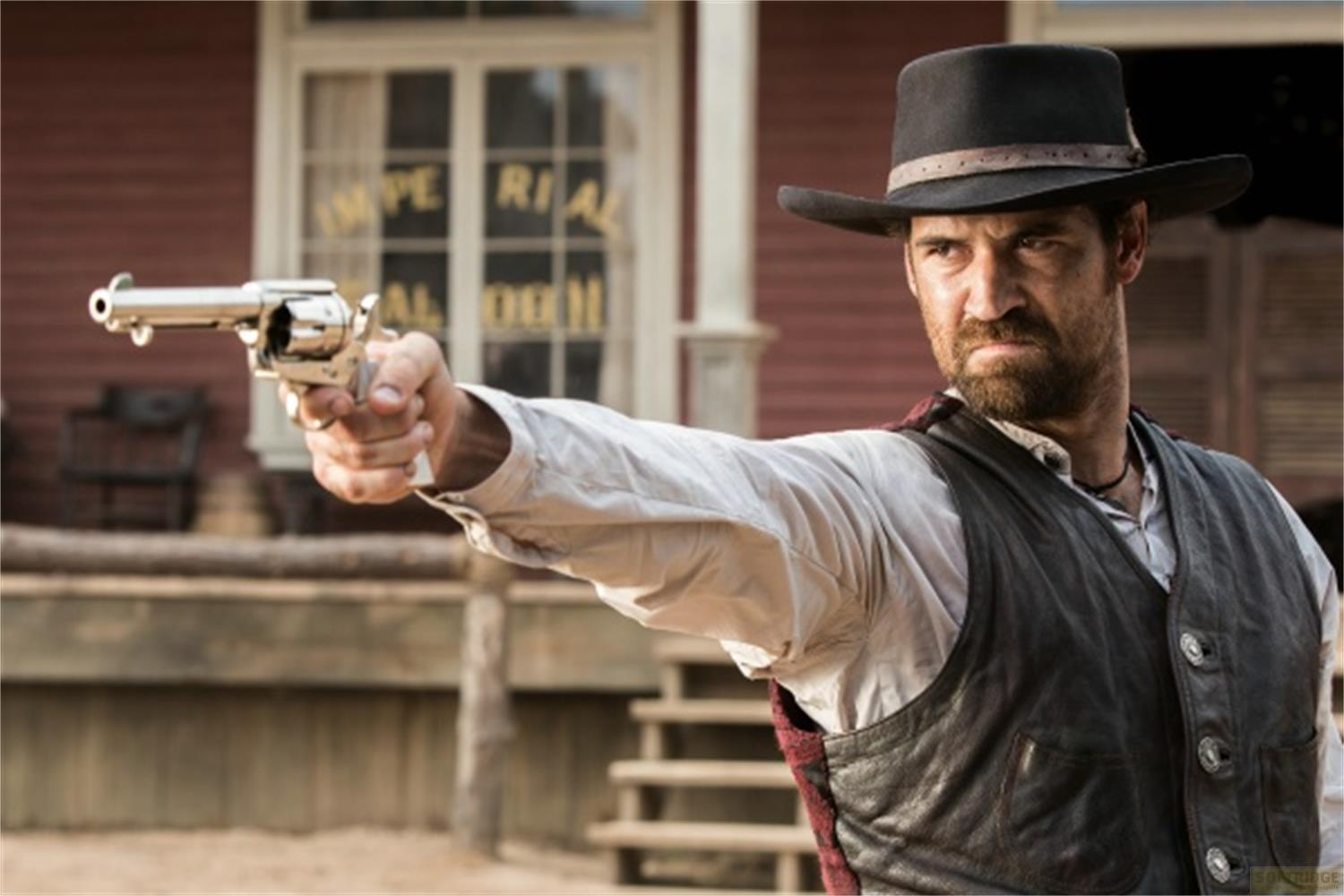 Homme au chapeau de cow-boy pointe un revolver devant un bâtiment rouge