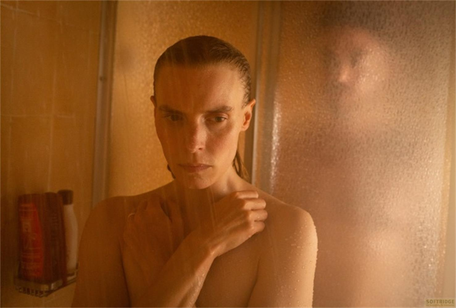 Una donna è sotto la doccia, l'acqua le cade addosso. Una seconda persona è visibile dietro una porta di vetro smerigliato.