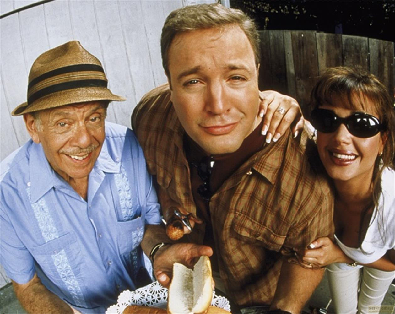 Tre persone sorridono in primo piano. Un uomo anziano indossa un cappello e una camicia blu.