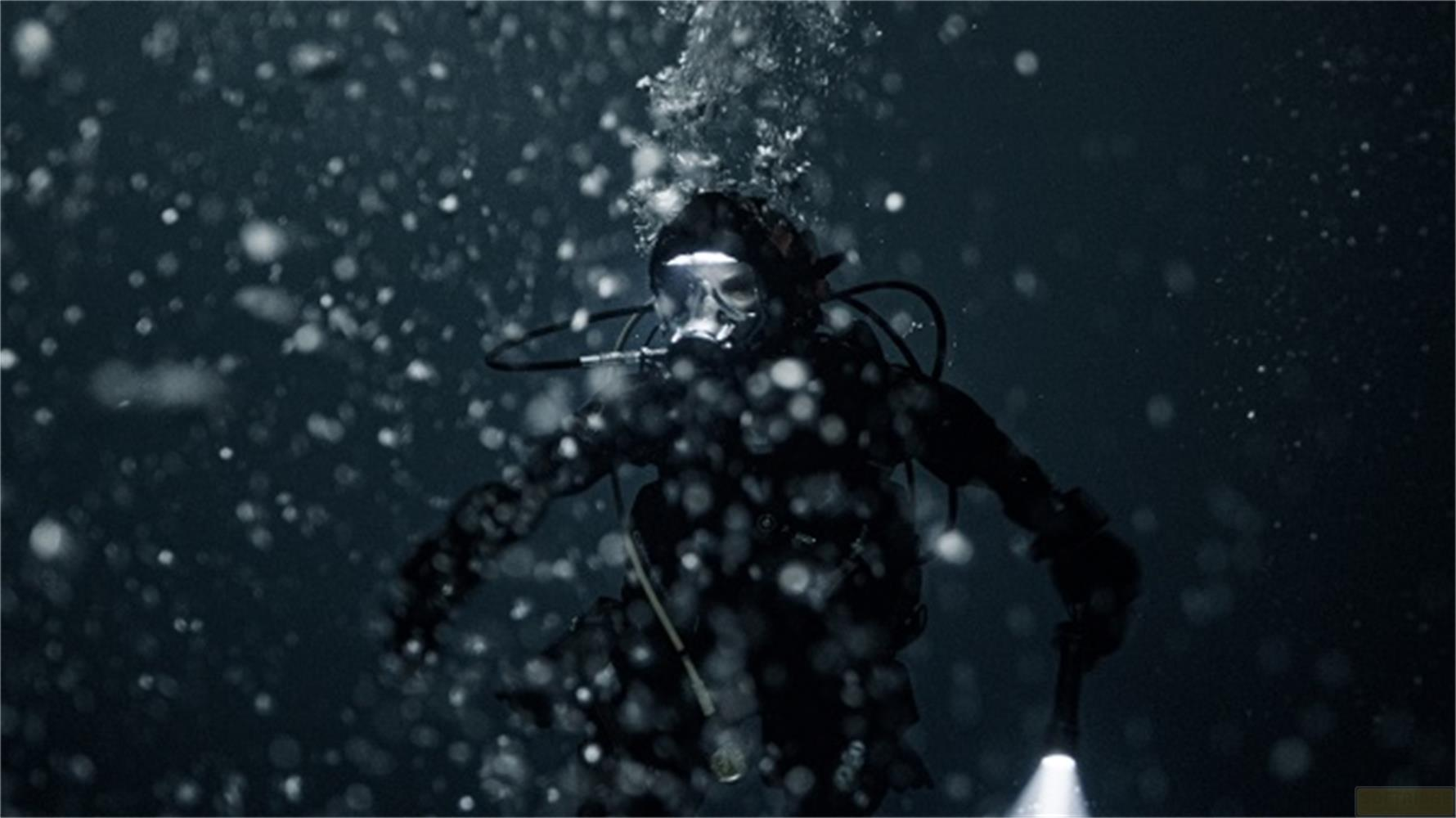 Un subacqueo in piena attrezzatura scende in acqua scura, illuminato da lampada frontale e torcia.